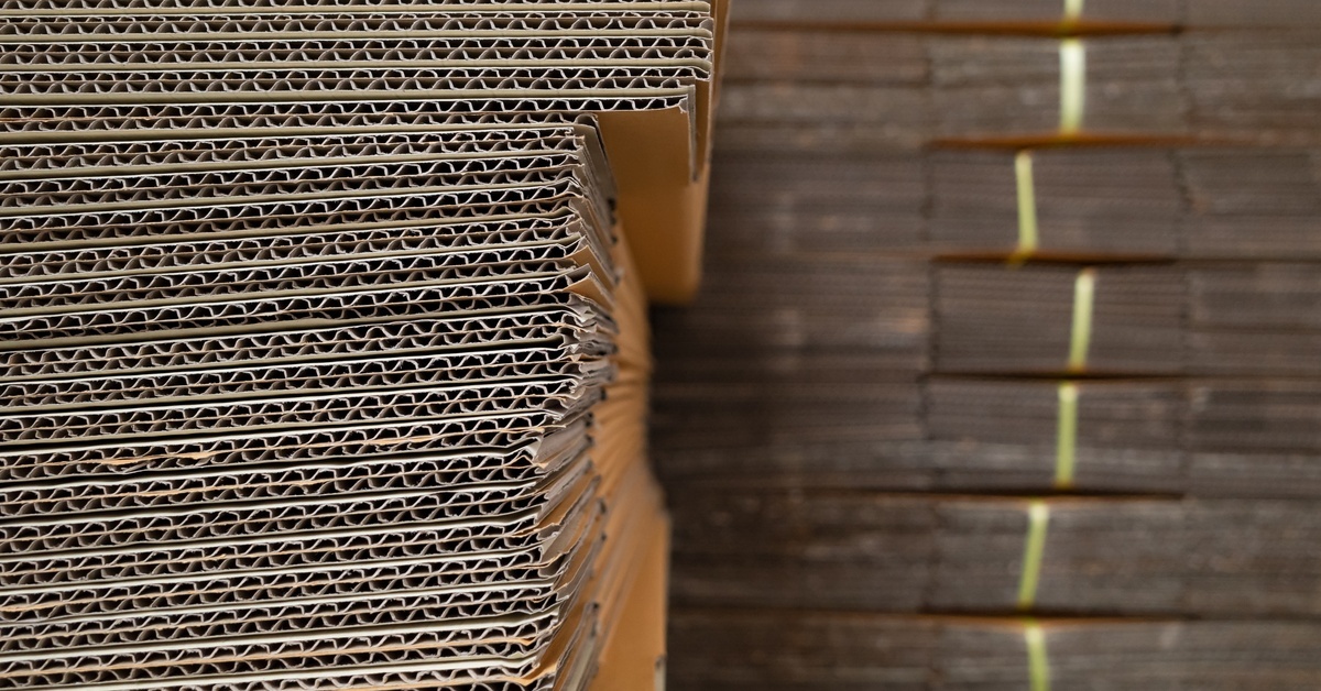 Corrugated cardboard containers sit in large stacks in a warehouse. Some of the boxes feature nylon ties.