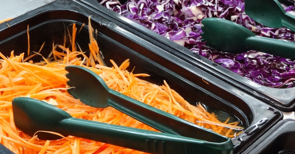 Two green plastic tongs lie in containers at a salad bar. One container has carrots, and another is full of red cabbage.