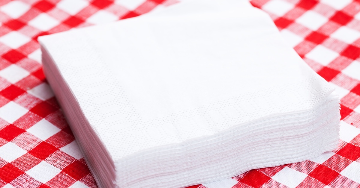 A stack of square paper napkins for drinks lies on a red and white gingham tablecloth in the center of the frame.