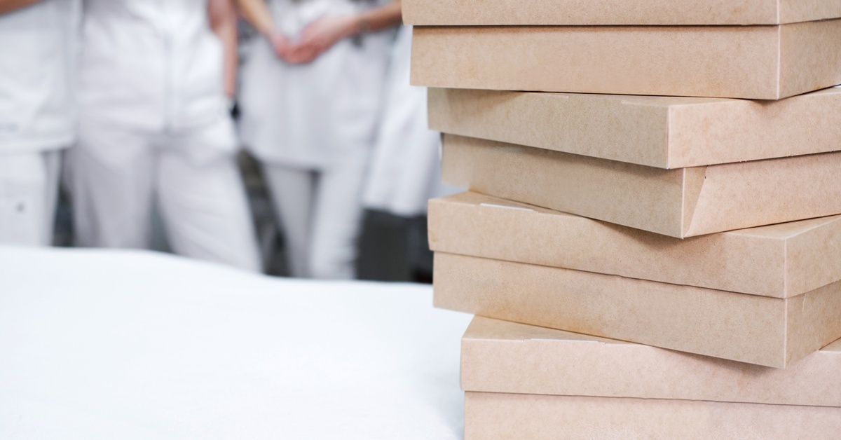 Several cardboard catering boxes sit stacked on a white counter, while people stand nearby in the background. Several cardboard catering boxes sit stacked on a white counter, while people stand nearby in the background.