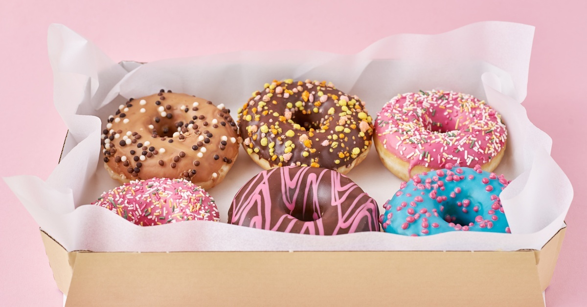 Multiple donuts of different colors sit on top of waxed paper inside a brown cardboard box. The box sits on a pink surface.