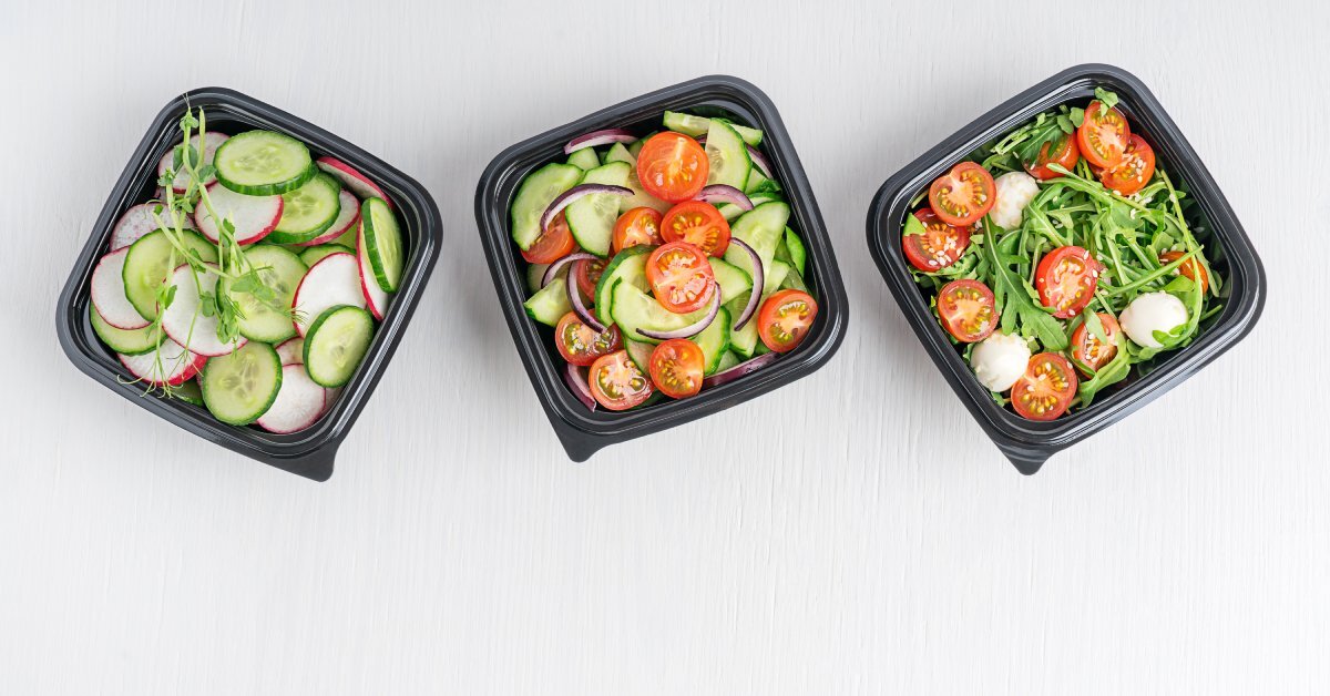 Overhead view of three black food containers sitting on a table. Each features a different vegetable salad.