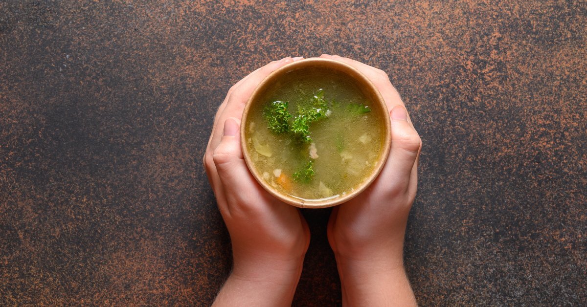 A person's hands hold a soup container as it sits on a table. Vegetable soup sits inside the container.