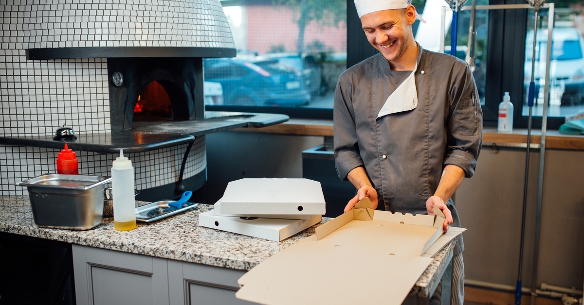 A pizzeria chef stands in the kitchen folding a pizza box together. Next to him is a large oven for the pizza.