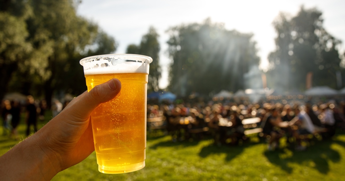 Close-up of a person's hand holding up a plastic cup of bright gold beer with a foamy head in front of an outdoor festival.