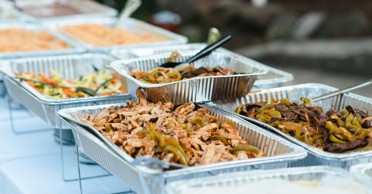 Different fajita ingredients sit inside several aluminum food trays on a long table at a catered event.