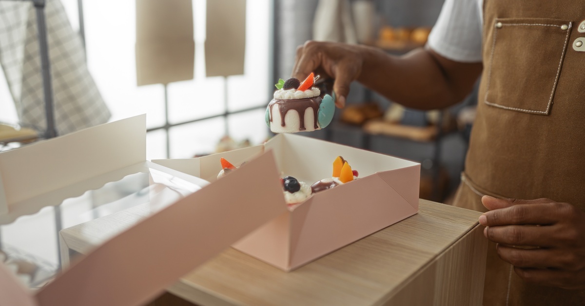 A baker's hands place small cakes into a bakery box that features a window. The baker wears a brown apron.