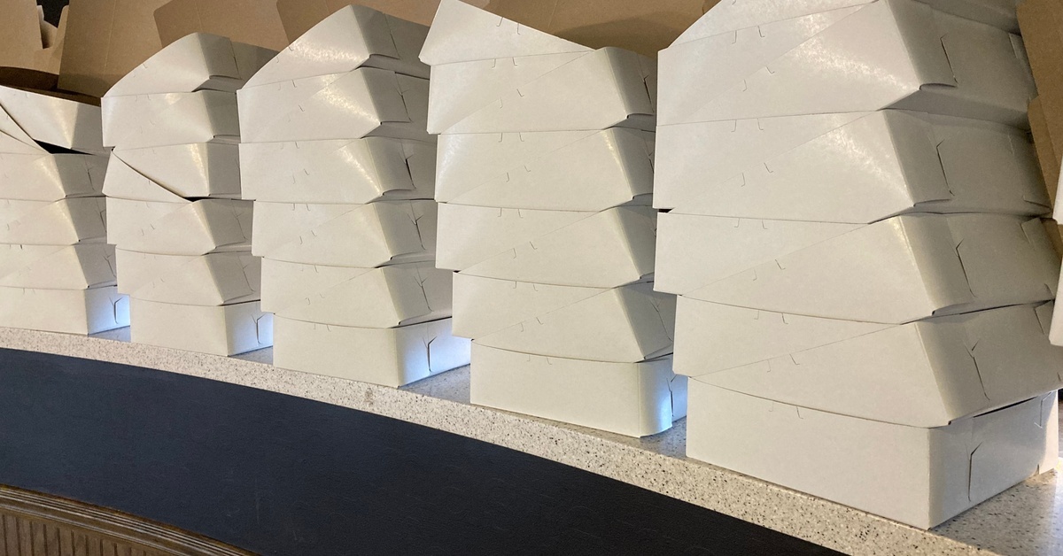 Several stacks of white bakery boxes sit alongside each other on a counter. The counter has a curved shape.