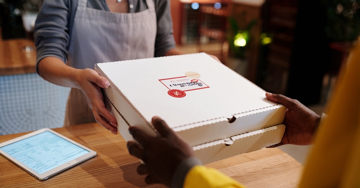A woman wearing an apron hands two pizza boxes off to another person. The top box features a red and white label.