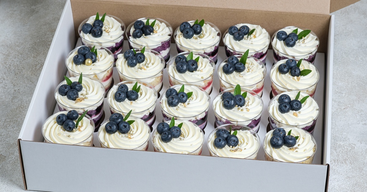 A large white cardboard box sits on a gray surface. It features several trifles in plastic cups inside.