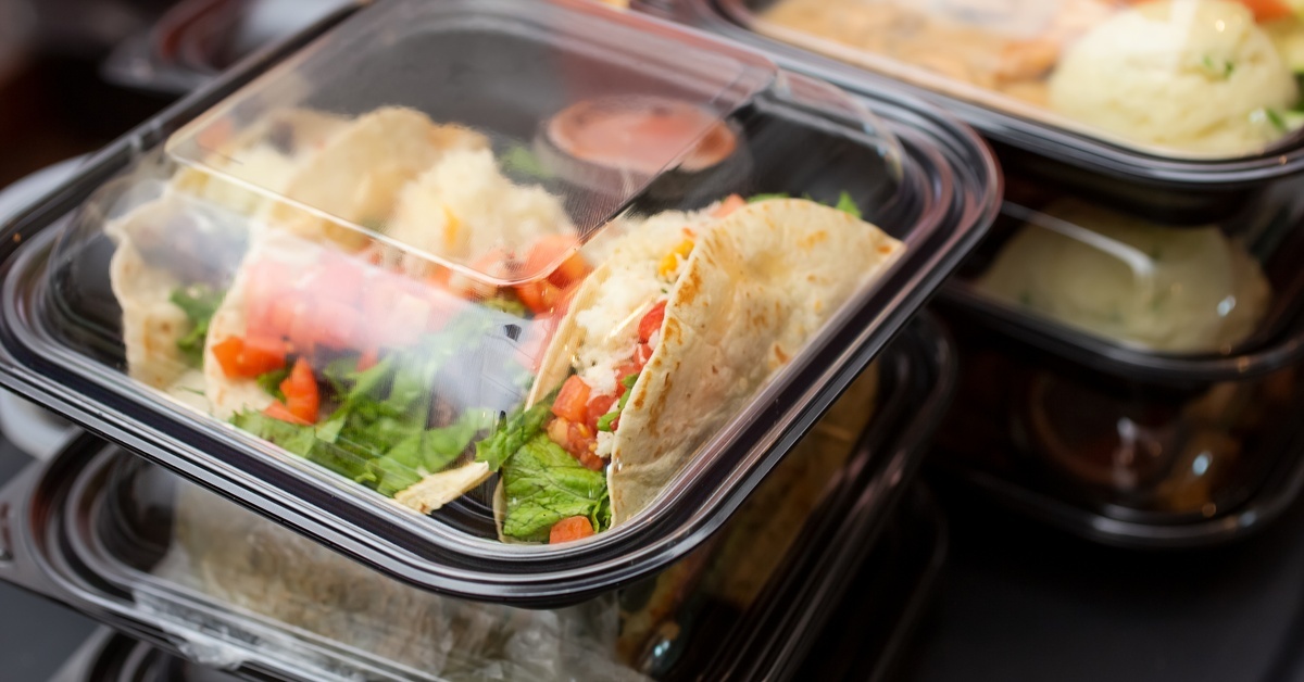 Several plastic food containers filled with food sit in stacks. One on top contains tacos and a container of sauce.