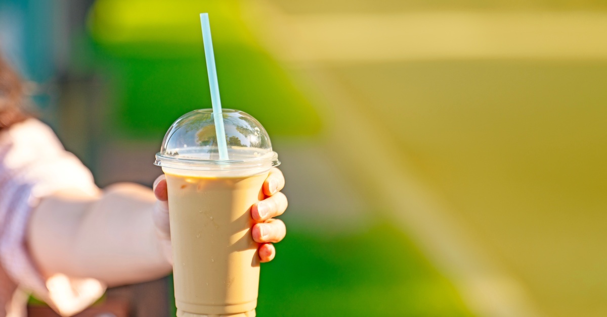 Close-up of a hand holding a tan-colored iced coffee in a plastic cup with a lid. The background is green and yellow