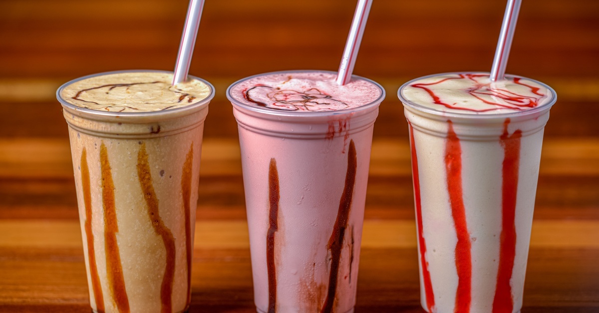 Three plastic cups featuring different milkshakes sit on a wooden counter. All three have straws in them.