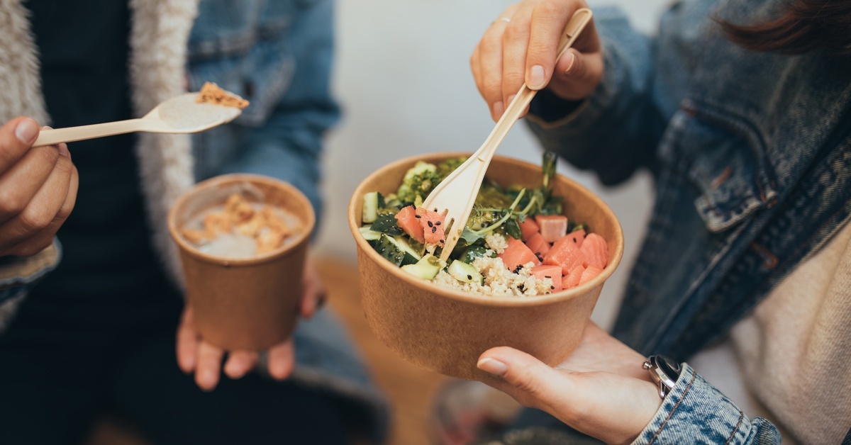 A close-up of two people holding cardboard takeout containers of soup and salad. They are using plastic utensils to eat from them.