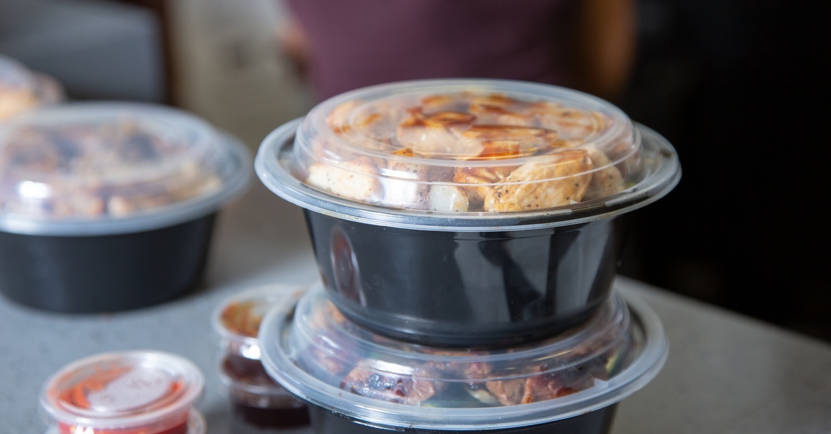 Black plastic to-go containers sit stacked up on a metal counter. Small containers of sauce sit next to them.
