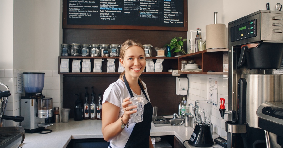 A barista in a coffee shop smiles as she holds a clear plastic cup with ice and water inside. She wears an apron.