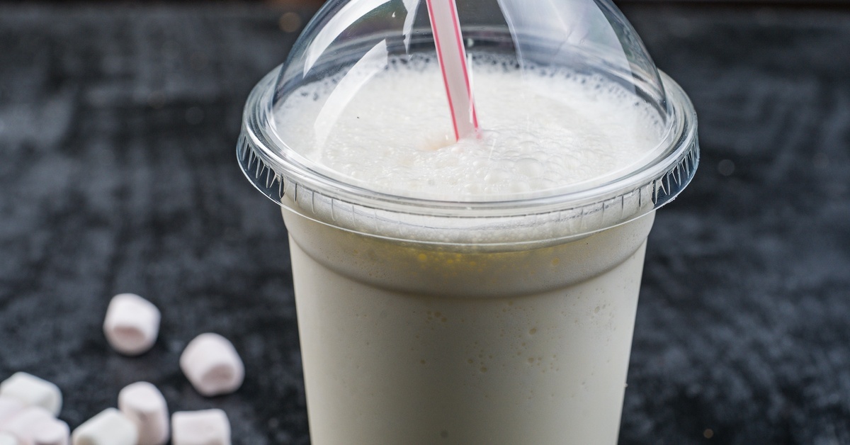 A vanilla milkshake sits inside a plastic cup with a domed lid on a counter. Several marshmallows sit nearby.