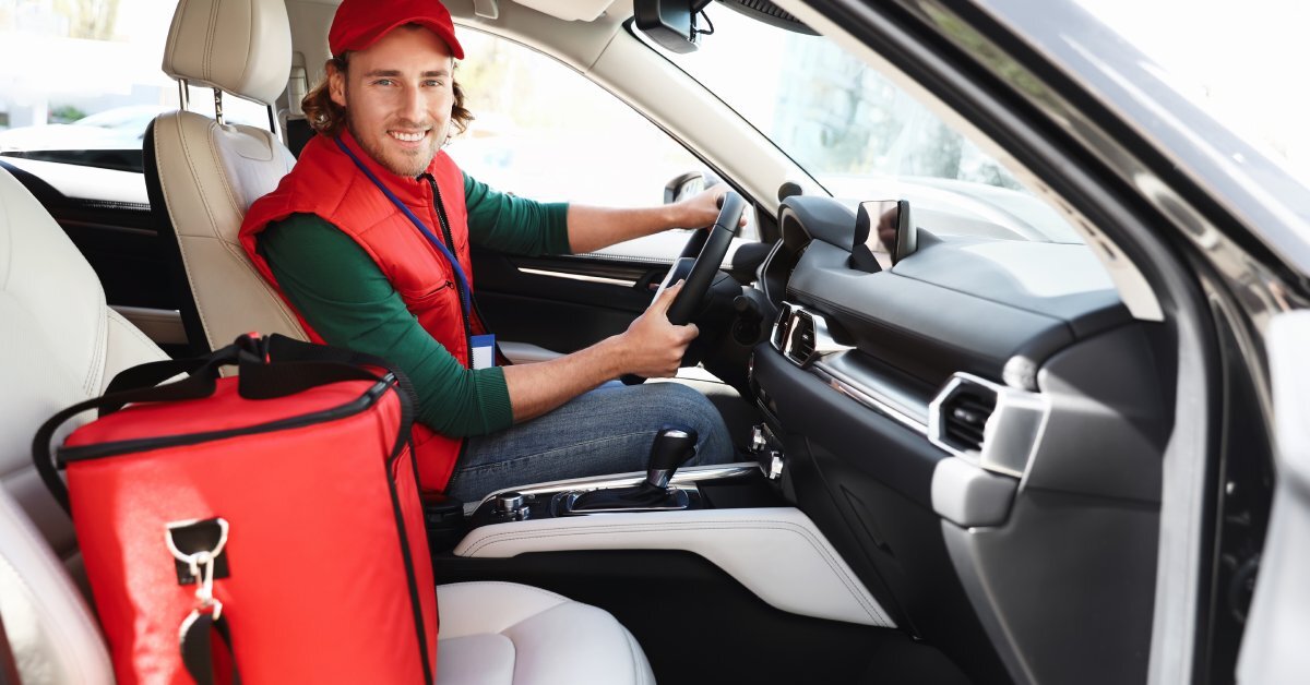 A man sits behind the wheel of his car with a red delivery bag next to him in the passenger seat. He wears a red cap.
