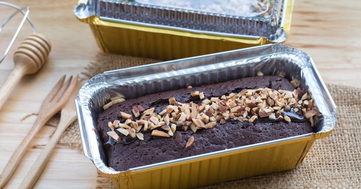 Two chocolate cakes with almond toppings sit inside aluminum foil containers on a table. One has a clear lid.