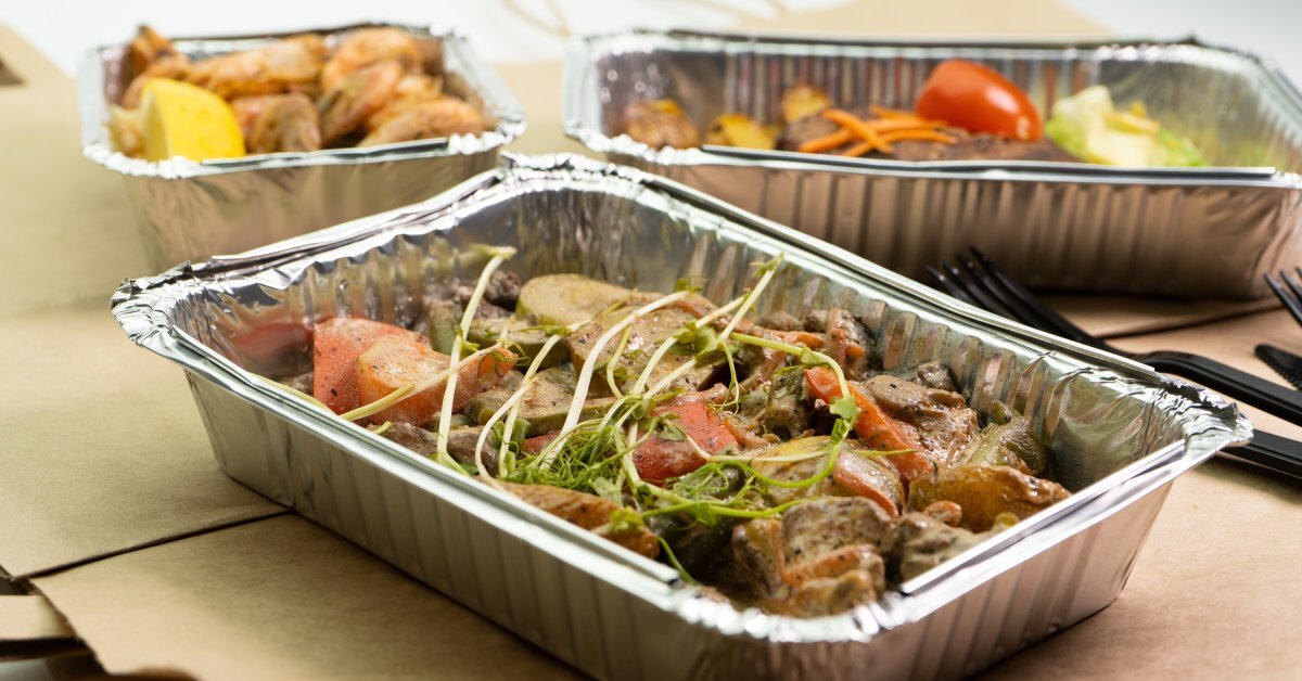 Three aluminum foil food containers sit on top of brown paper bags. They feature a variety of items, including roast beef.